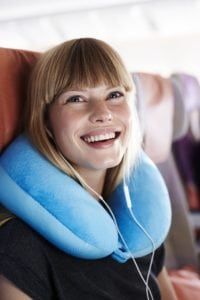 Young female airplane passenger with headphones smiling while wearing a travel pillow and sitting up straight